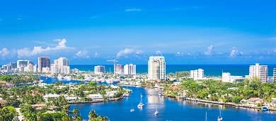 Scenic view of Fort Lauderdale, Florida, featuring a waterway lined with yachts, palm trees, and luxury homes, with a backdrop of modern high-rise buildings and the Atlantic Ocean under a bright blue sky.